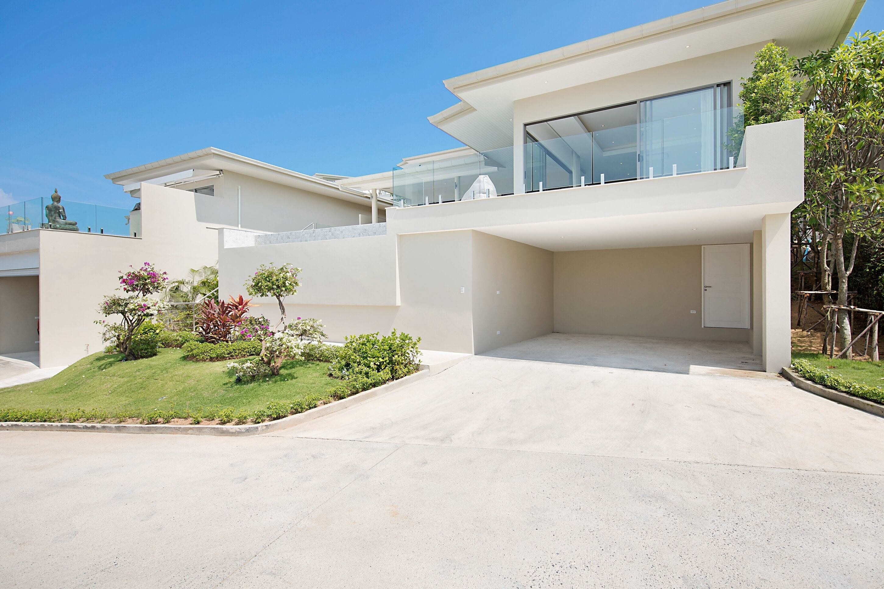 A modern house with a spacious driveway, a small garden with various plants, and a large glass window on the upper floor.