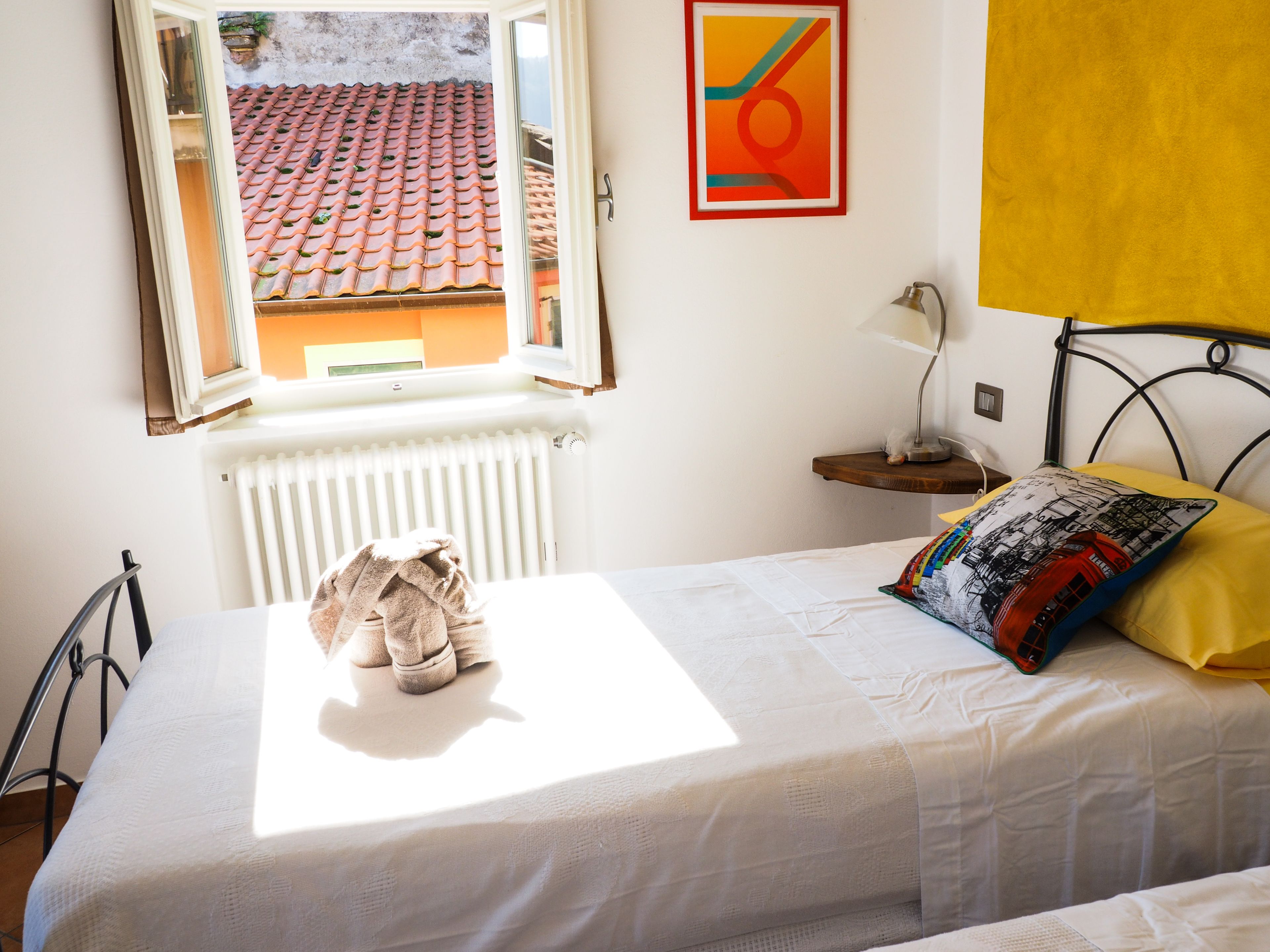 Quarto com cama, janela com vista aberta, radiador, mesa de cabeceira com abajur e travesseiro colorido.