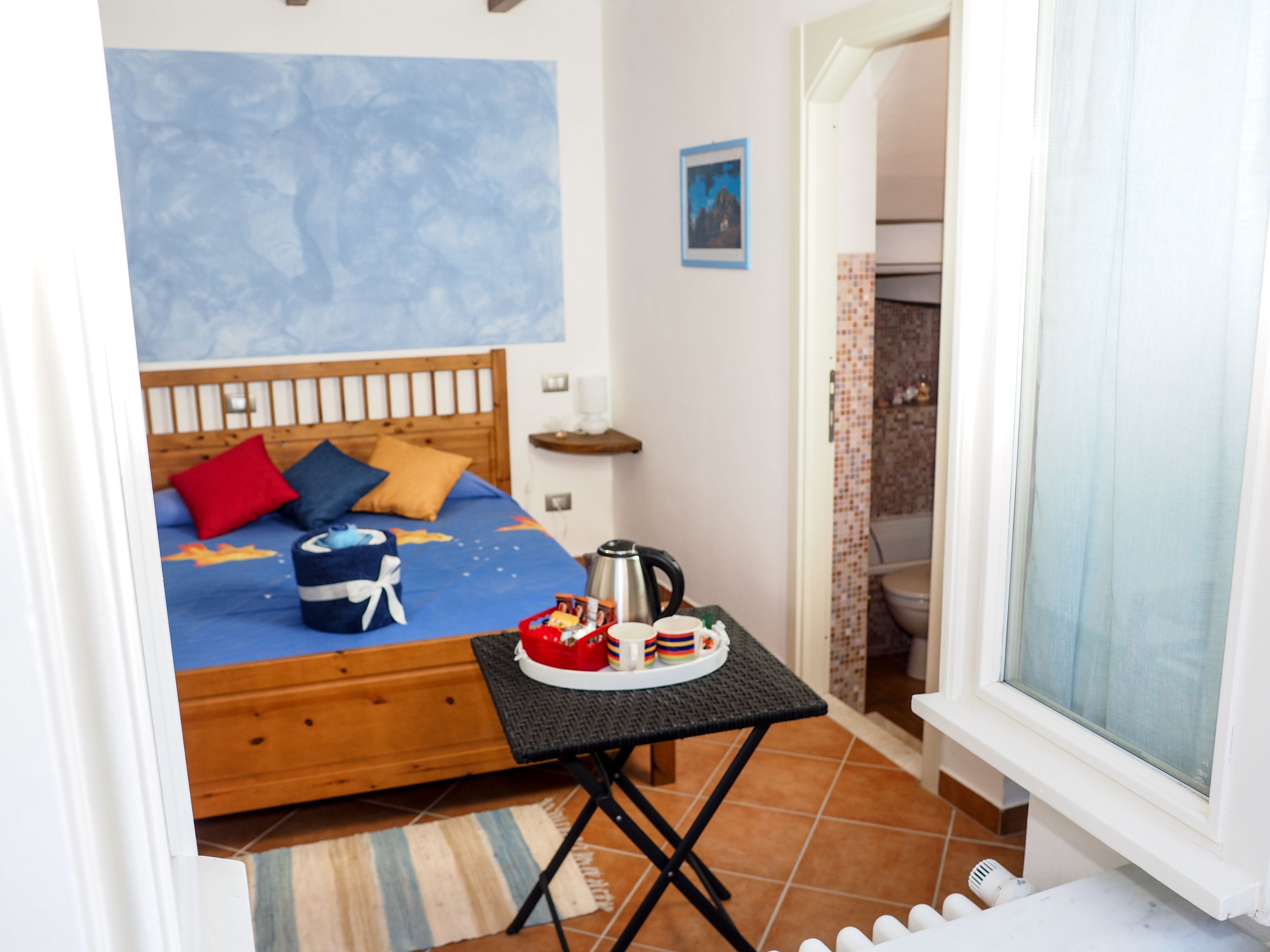 A bedroom with a bed, a small table with a kettle, and a window.
