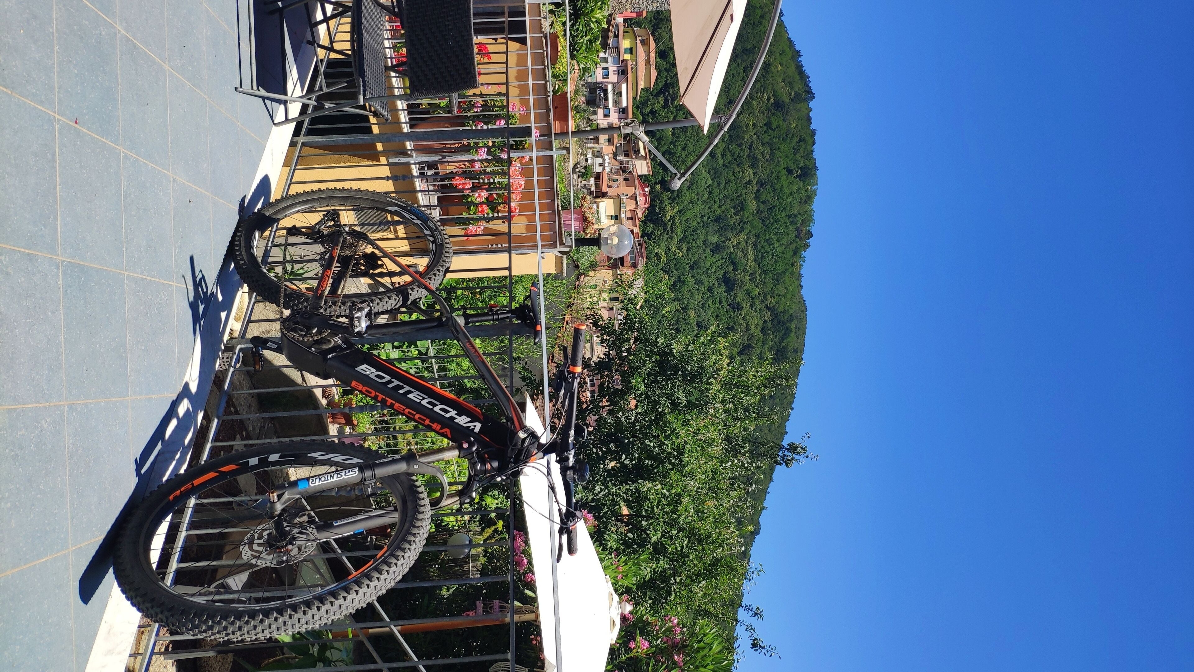Uma bicicleta estacionada em um suporte de metal, com vista para uma área residencial e uma colina ao fundo.