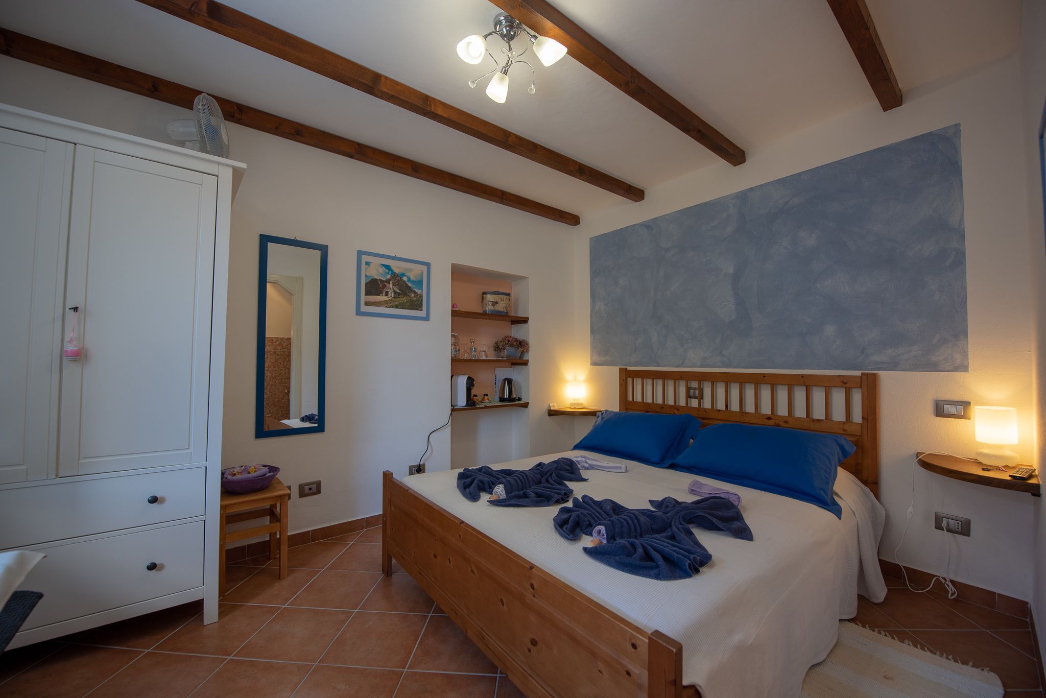 A bedroom with a wooden bed, a white wardrobe, a small table, and a wall-mounted shelf.