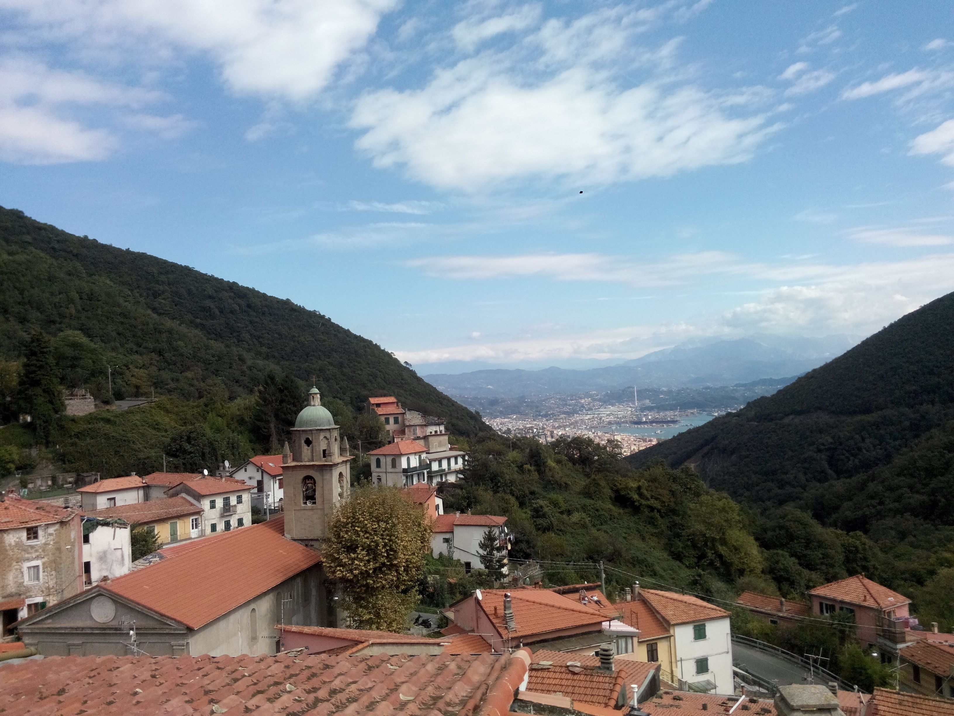 A town with a prominent church featuring a bell tower and a dome, surrounded by terraced houses with red-tiled roofs, set against a backdrop of hills and a clear sky.