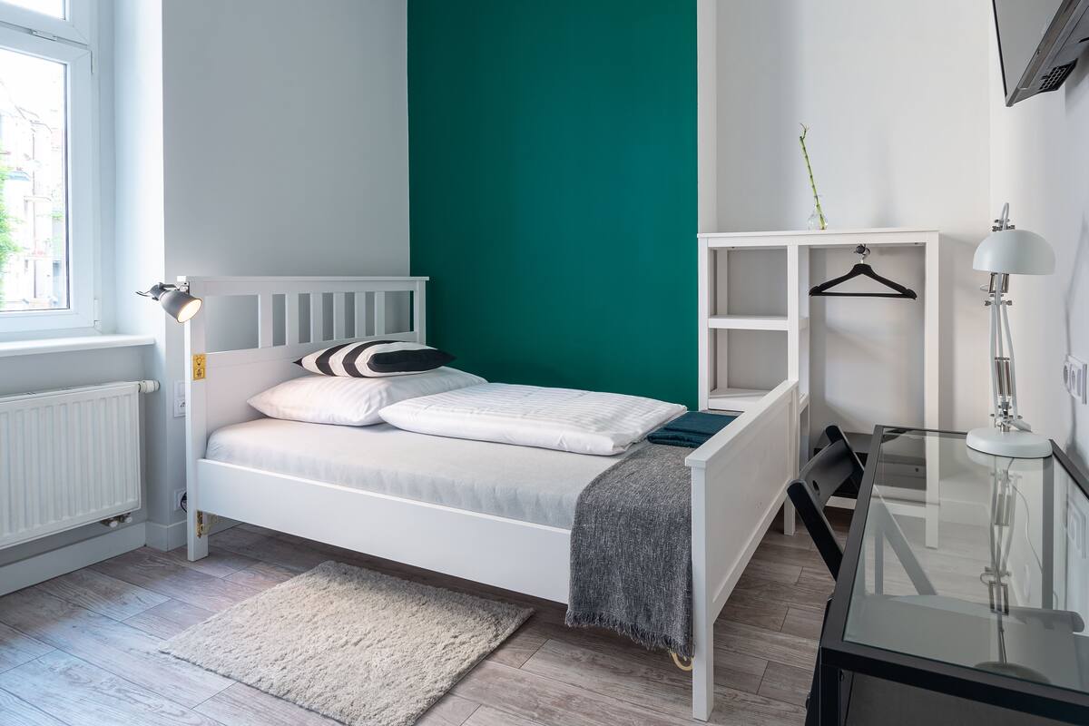 A bedroom with a white bed, teal accent wall, white radiator, and a desk with a lamp.