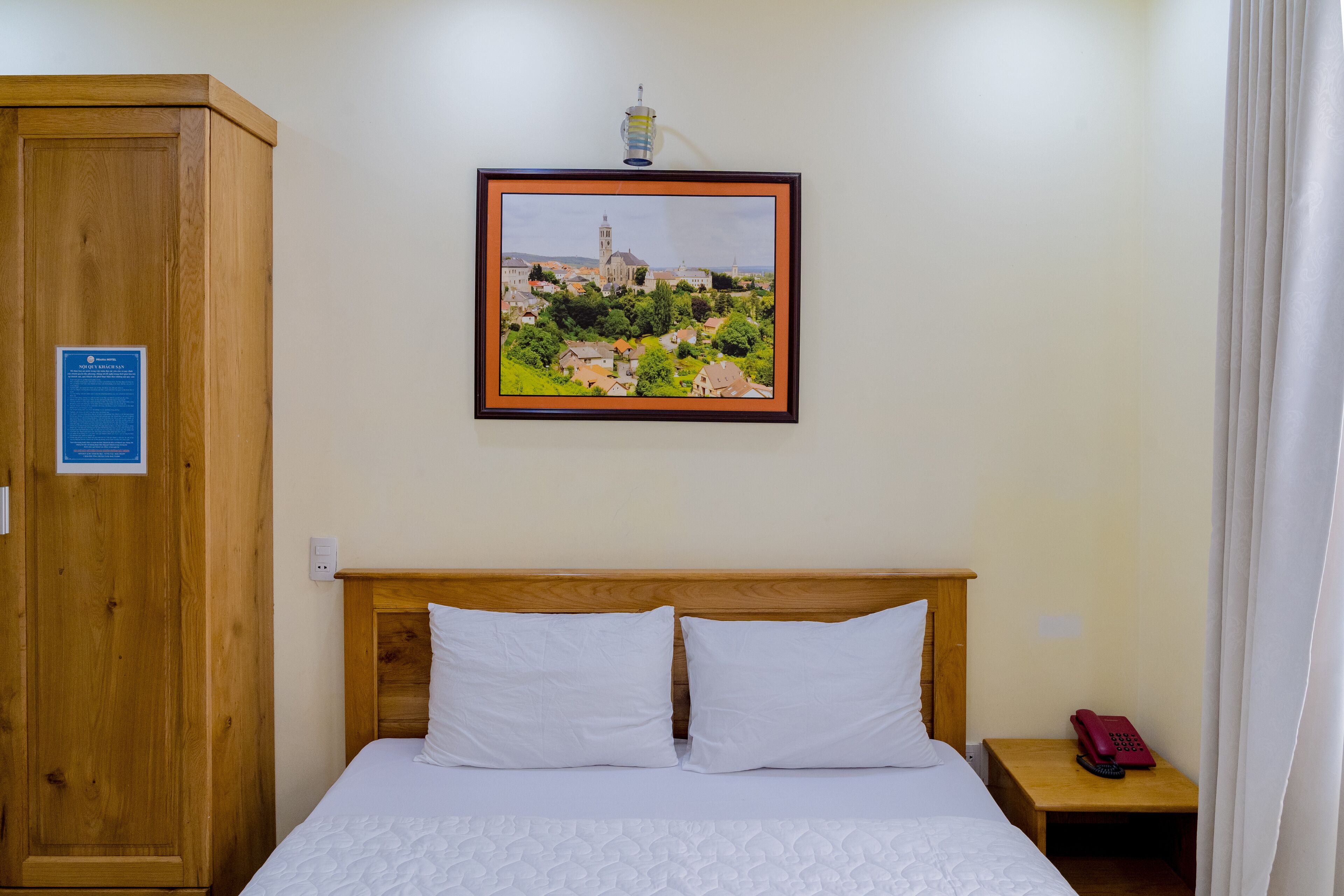 Una habitación de hotel con cabecera de madera, cama con ropa de cama blanca, un cuadro con vista urbana en la pared, un armario de madera y una mesita de noche con teléfono.