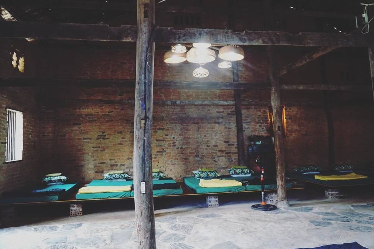 Ein rustikaler Raum mit Backsteinwänden, Holzbalken und niedriger Decke. Vier Betten stehen entlang der Wand, jedes mit einer grünen Tagesdecke und Kissen. Eine Chandelier hängt von der Decke, und ein kleiner Tisch mit einer Lampe ist auf der rechten Seite zu sehen.