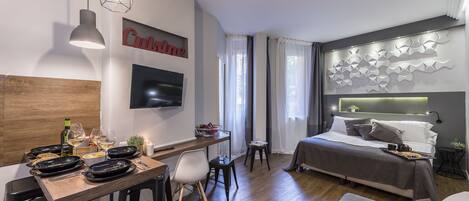 A modern hotel room with a wooden dining table, a bed with a headboard, a TV mounted on the wall, and a decorative wall piece.