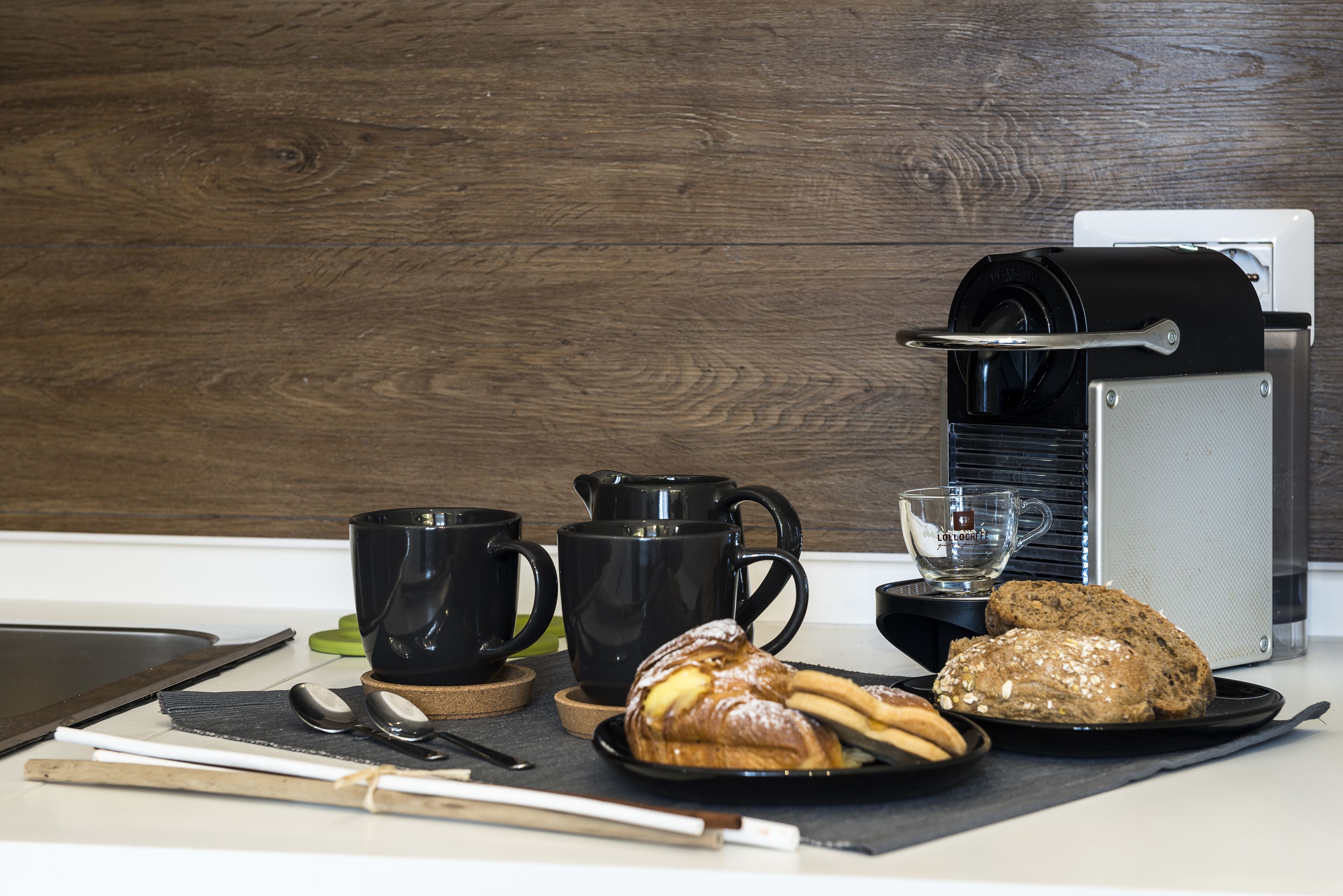 A kitchen counter with a coffee maker, black mugs, a glass, and pastries.