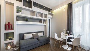 A modern living room with a grey sofa, white chairs, and a dining table. Shelves with decorative items and a clear side table with a candle are visible. - Z-ArchSuiTes (Rome)