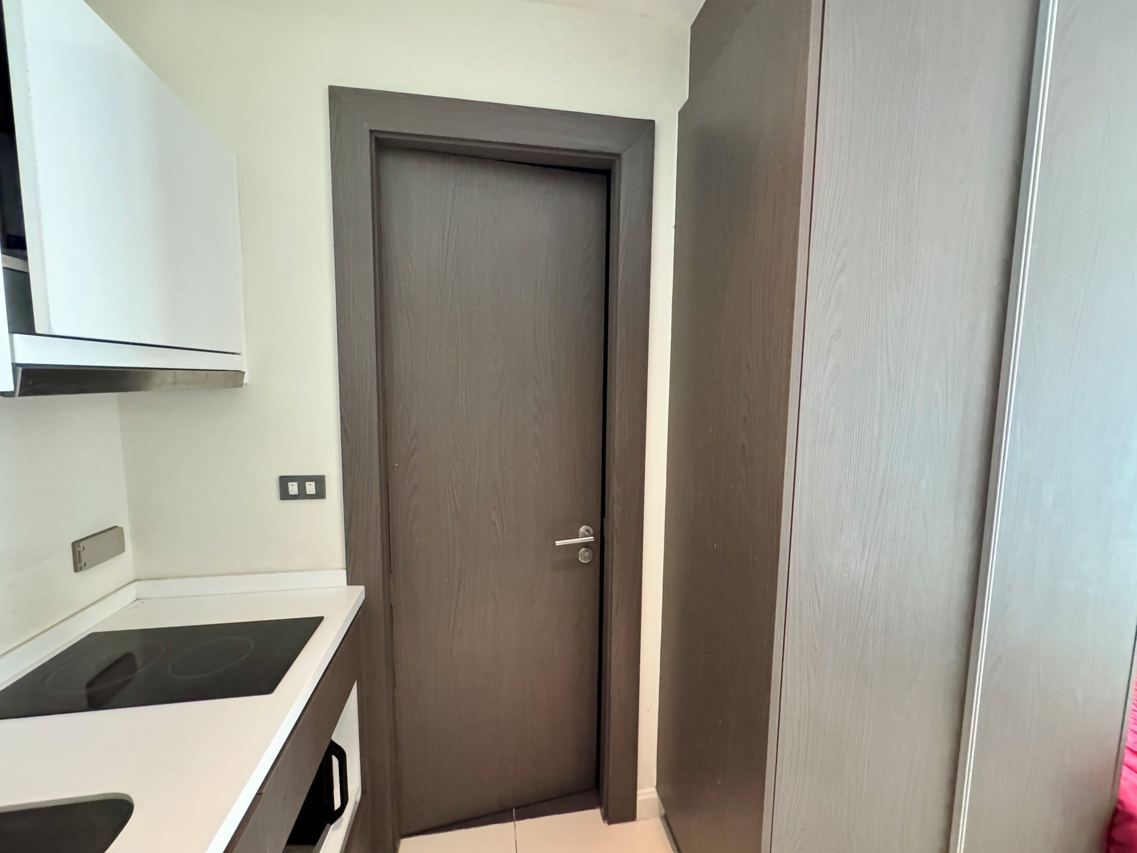 Eine kleine Küche mit weißer Arbeitsplatte, einer dunklen Holztür und einem weißen Schrank darüber.