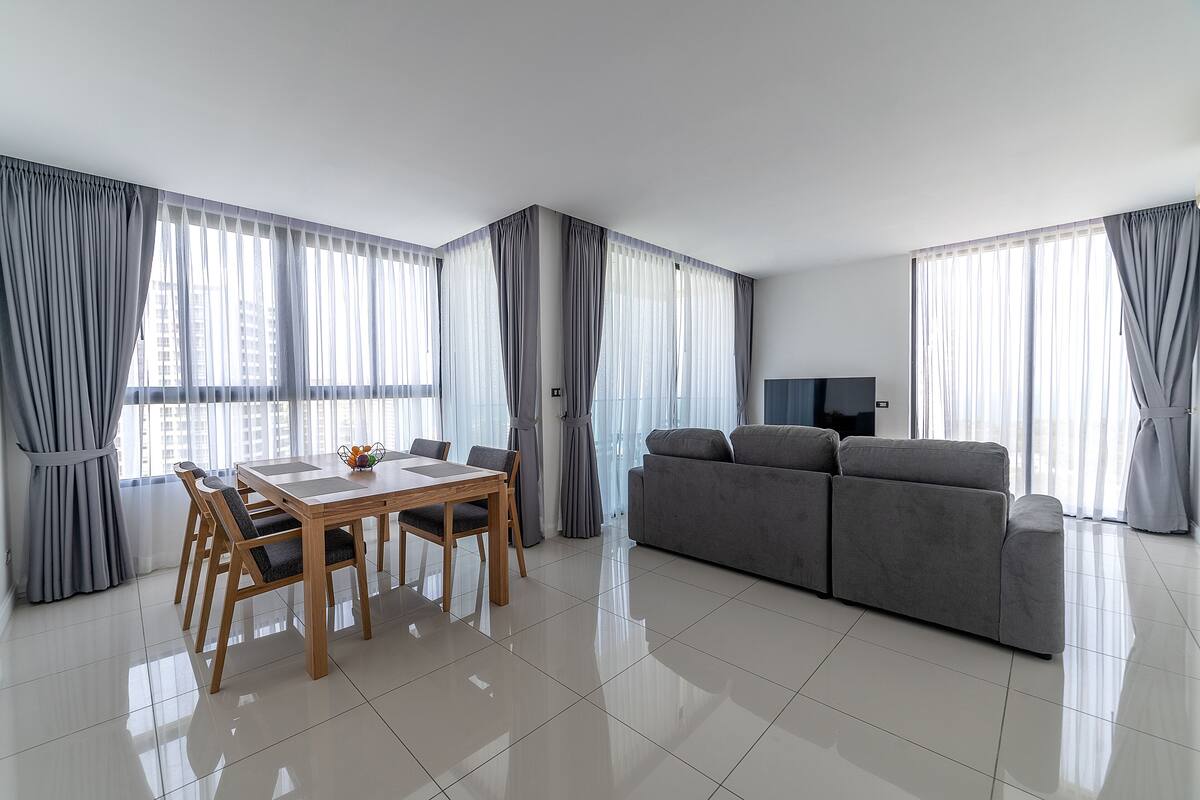 A modern living room with a dining area, grey curtains, a grey sofa, and a flat-screen TV.
