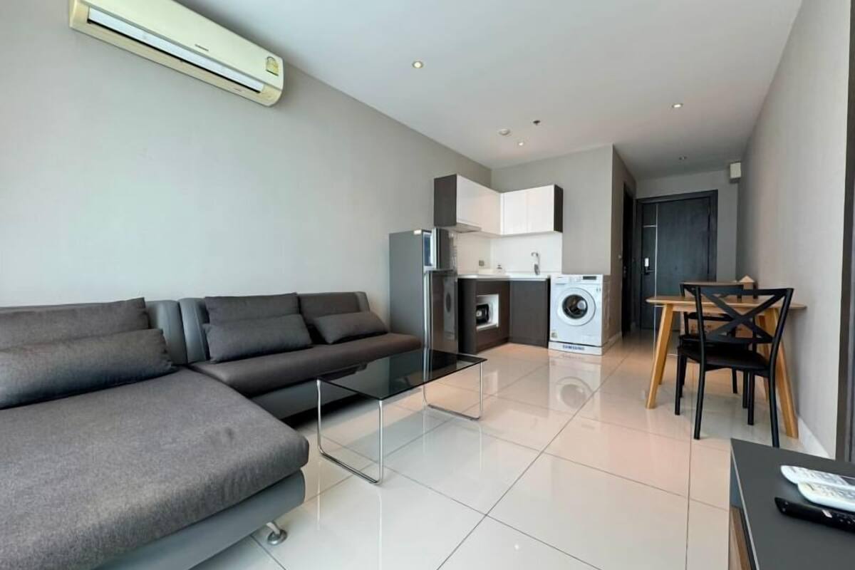 A modern living room with a grey sofa, a glass coffee table, a washing machine, and a dining area with wooden chairs.