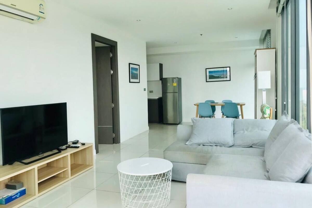A modern living room with a flat-screen TV, a grey sofa, a small white coffee table, and a kitchen area in the background.