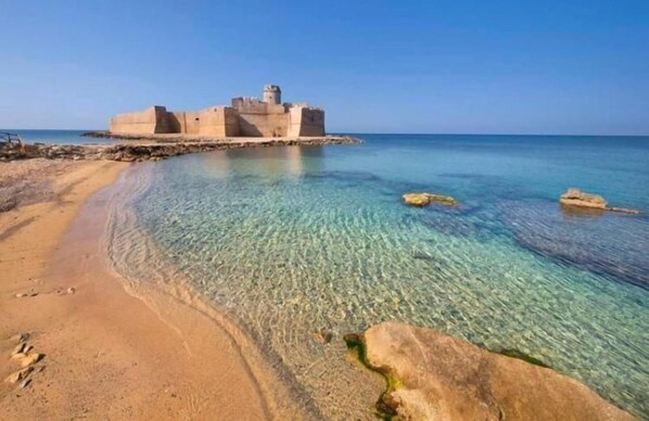Una playa costera con agua cristalina, una orilla arenosa y una fortificación histórica al fondo.
