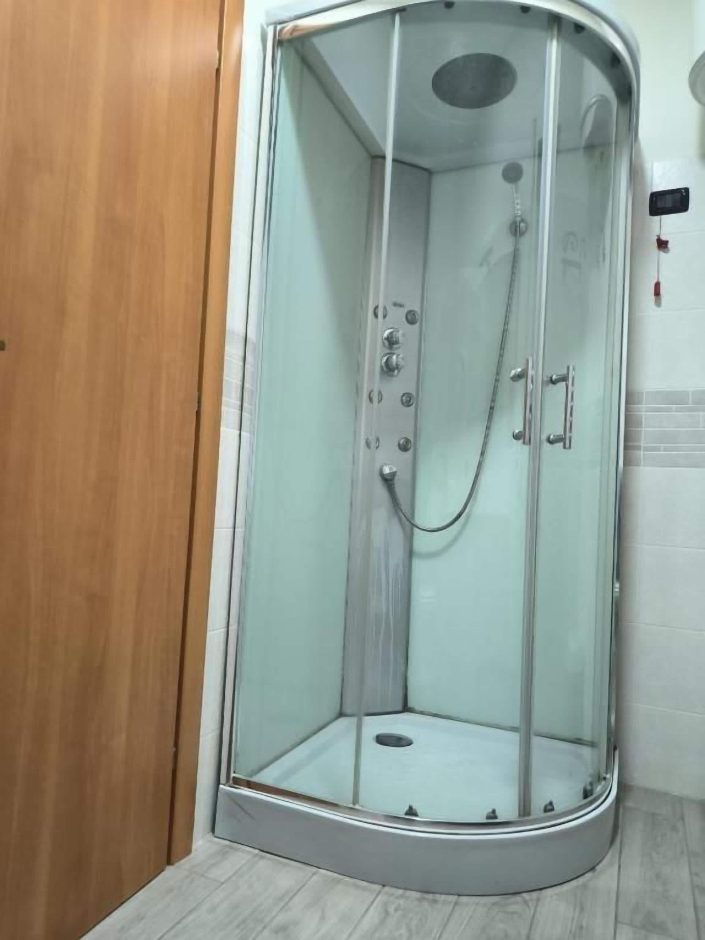 A glass-enclosed shower stall with a control panel, a handheld showerhead, and a handheld vacuum.