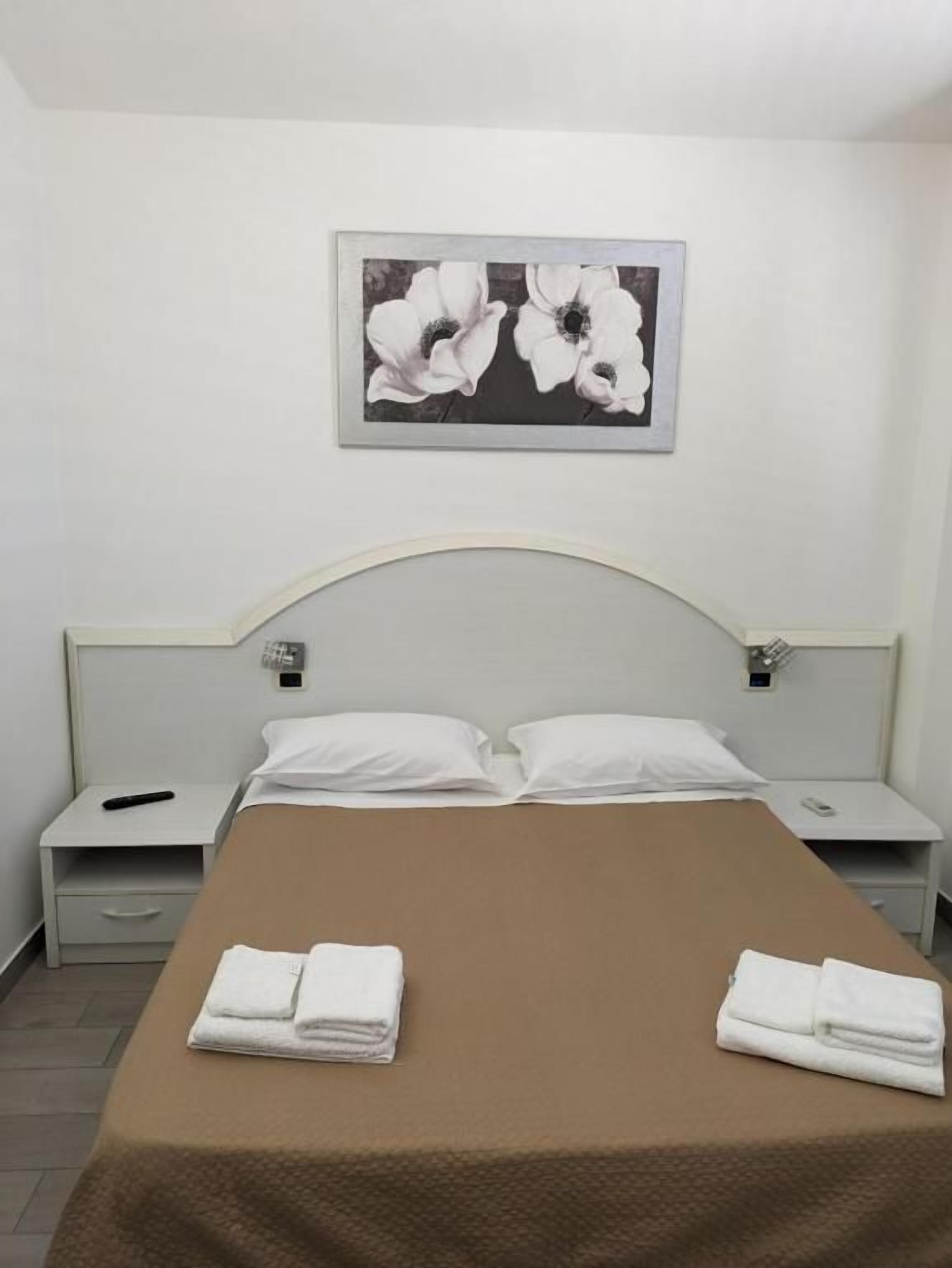 A hotel room with a bed, two pillows, a nightstand, a remote control, and a folded towel on the bed.