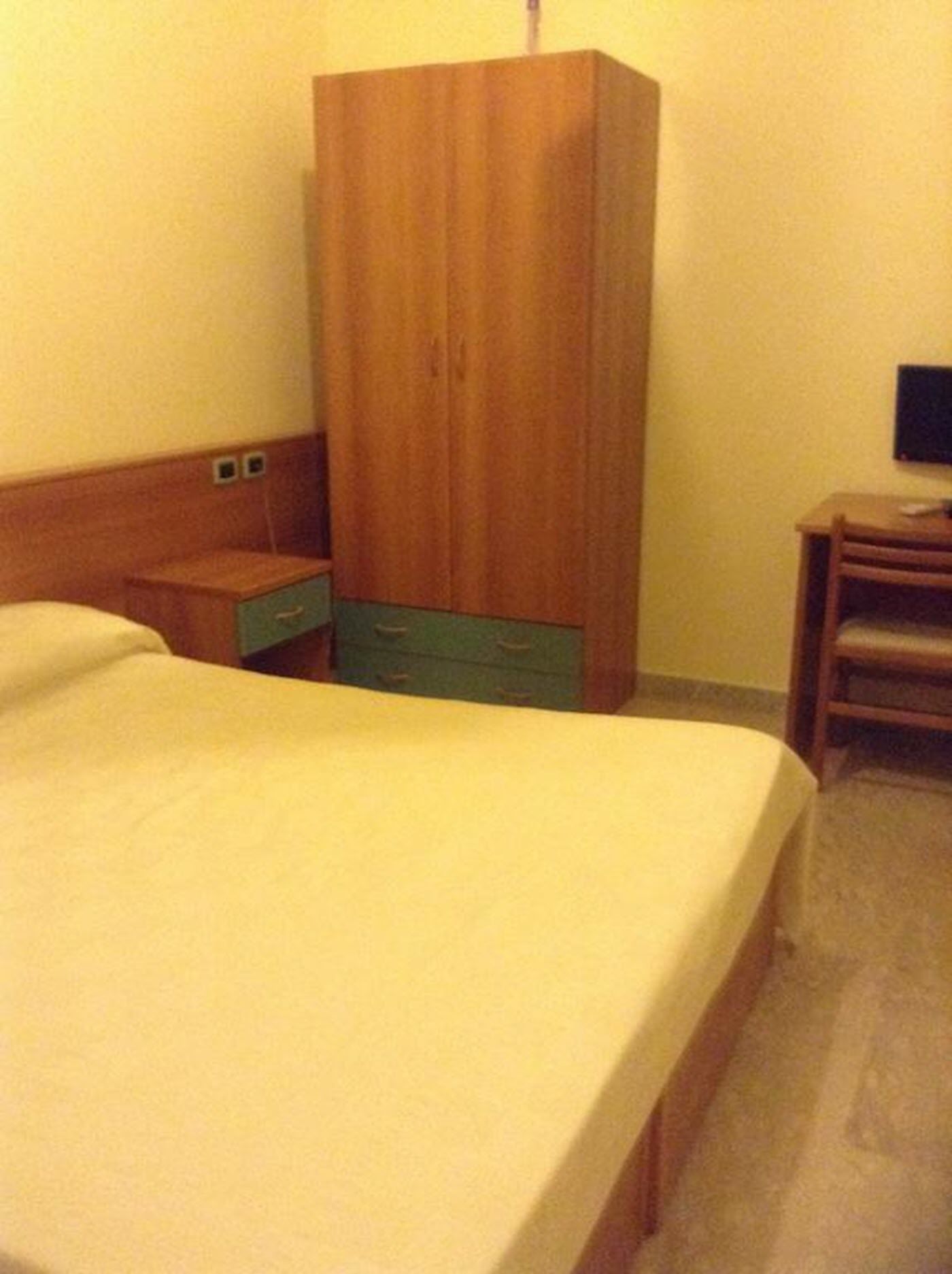 A hotel room with a bed, a wooden wardrobe, a nightstand, and a desk.