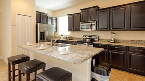 A modern kitchen with dark cabinetry, a granite countertop, and stainless steel appliances. There are three bar stools and a trash can.