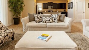 A living room with a beige sofa, a coffee table with a magazine, a patterned armchair, and a large potted plant.
