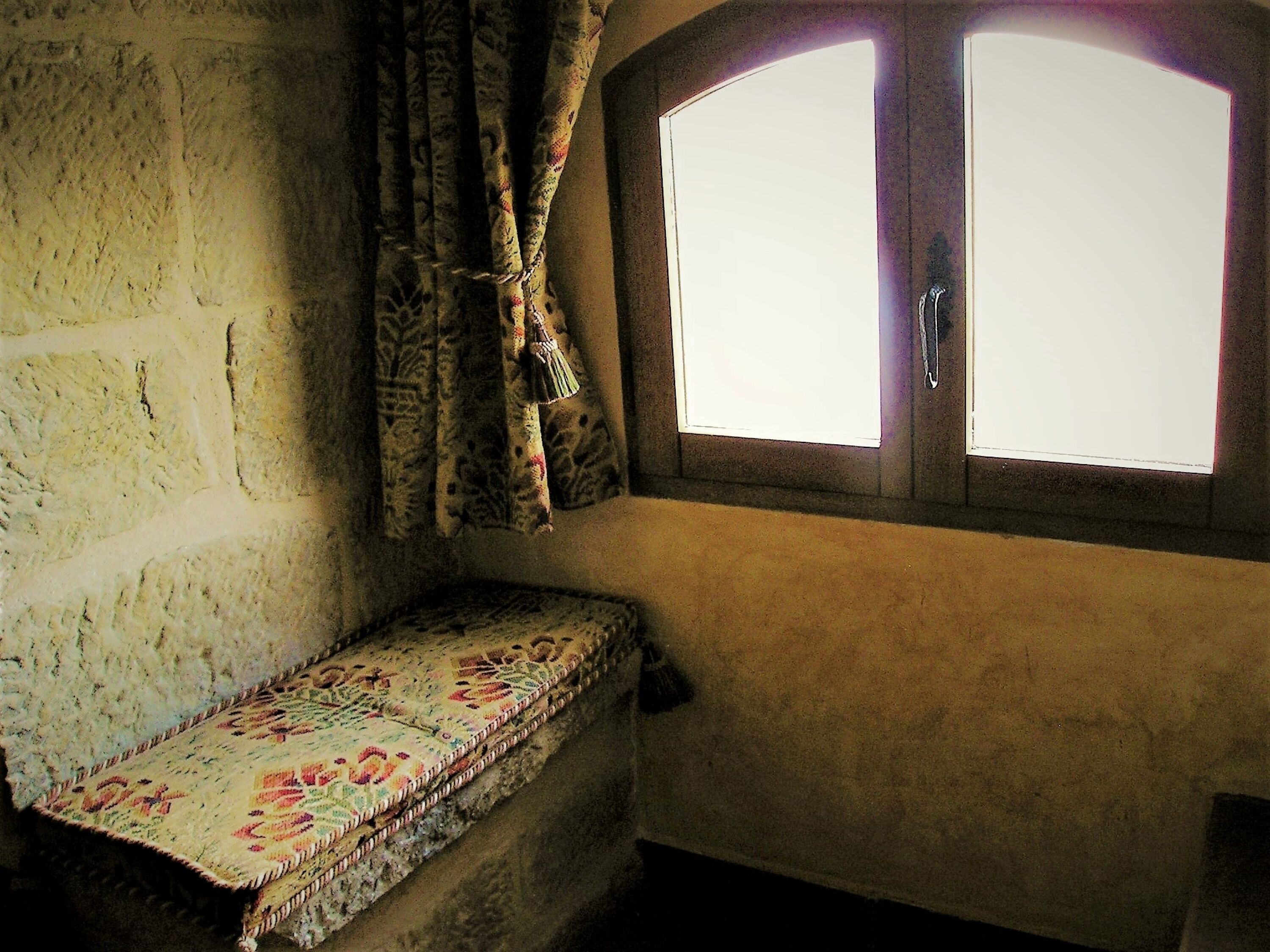 Una habitación con pared de piedra, un banco con cojín estampado y una ventana de dos cristales.