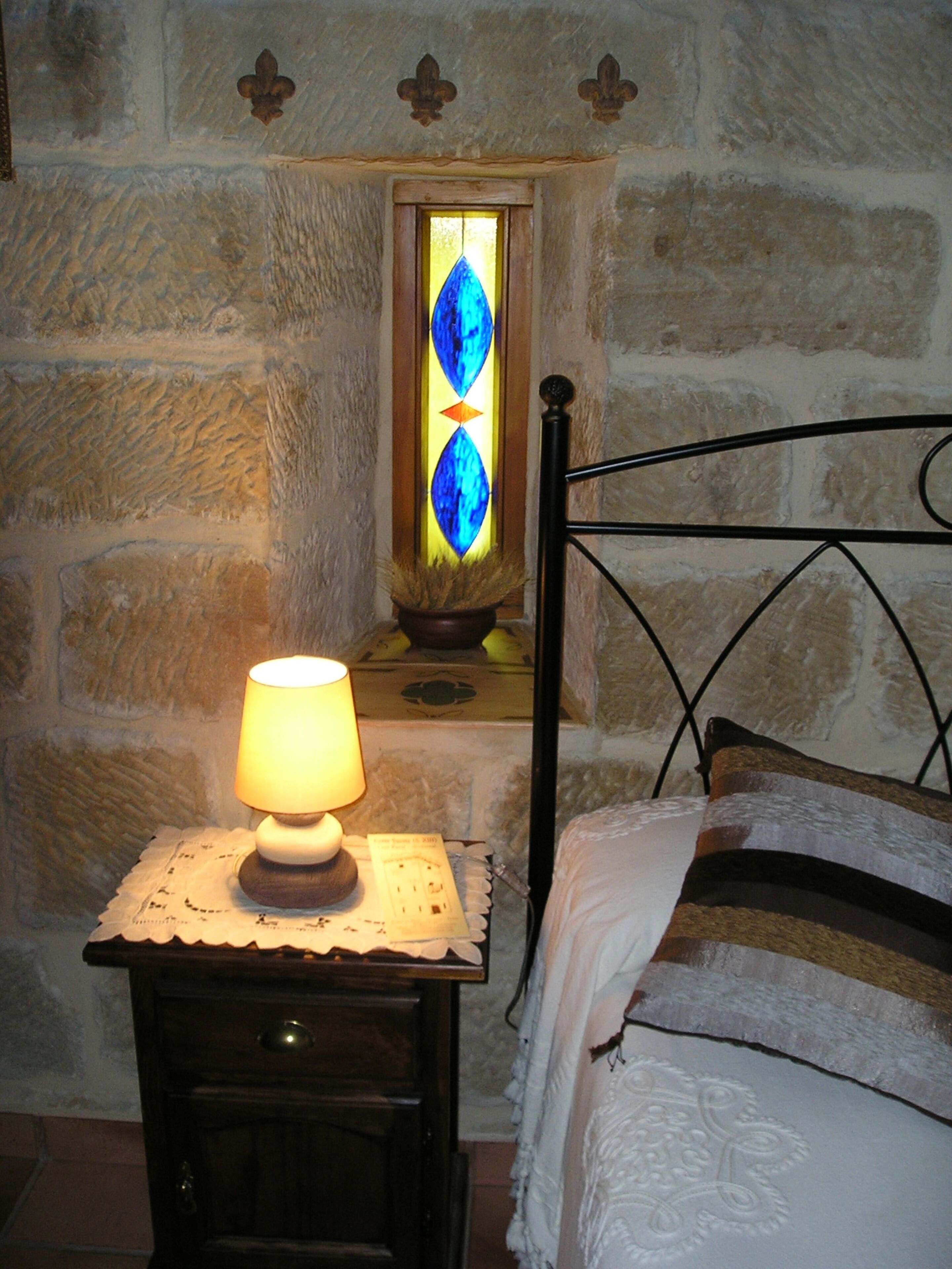 A bedroom with a stone wall, a bed with white and brown bedding, a bedside table with a lamp, and a stained glass window.