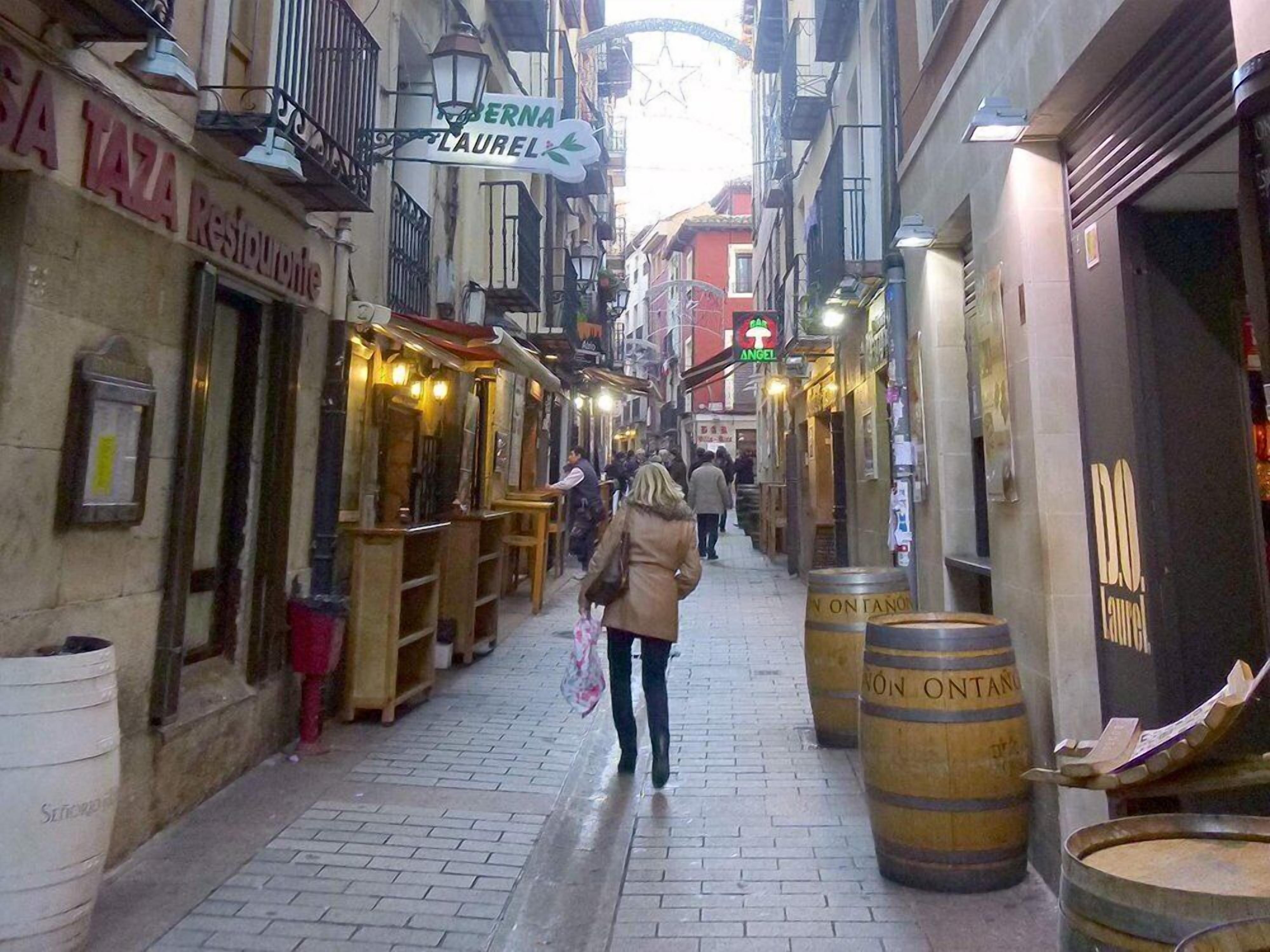 A narrow street lined with shops and restaurants, including 'SA TAZA Restaurant' and 'BERNA LAUREL'.