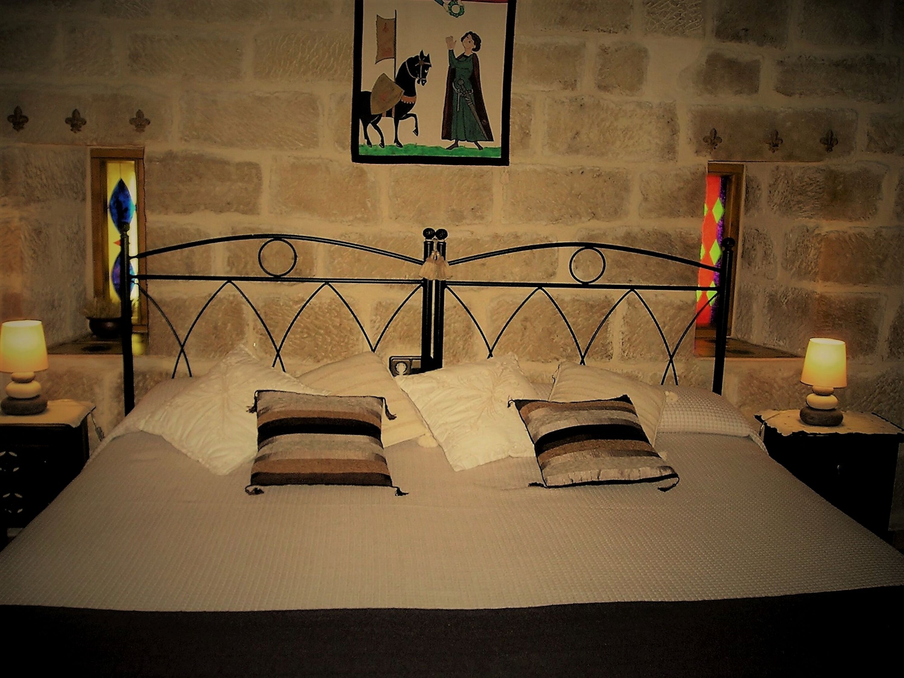 Una cama con cabecera de metal, dos mesitas de noche con lámparas y una ventana con vidrieras.