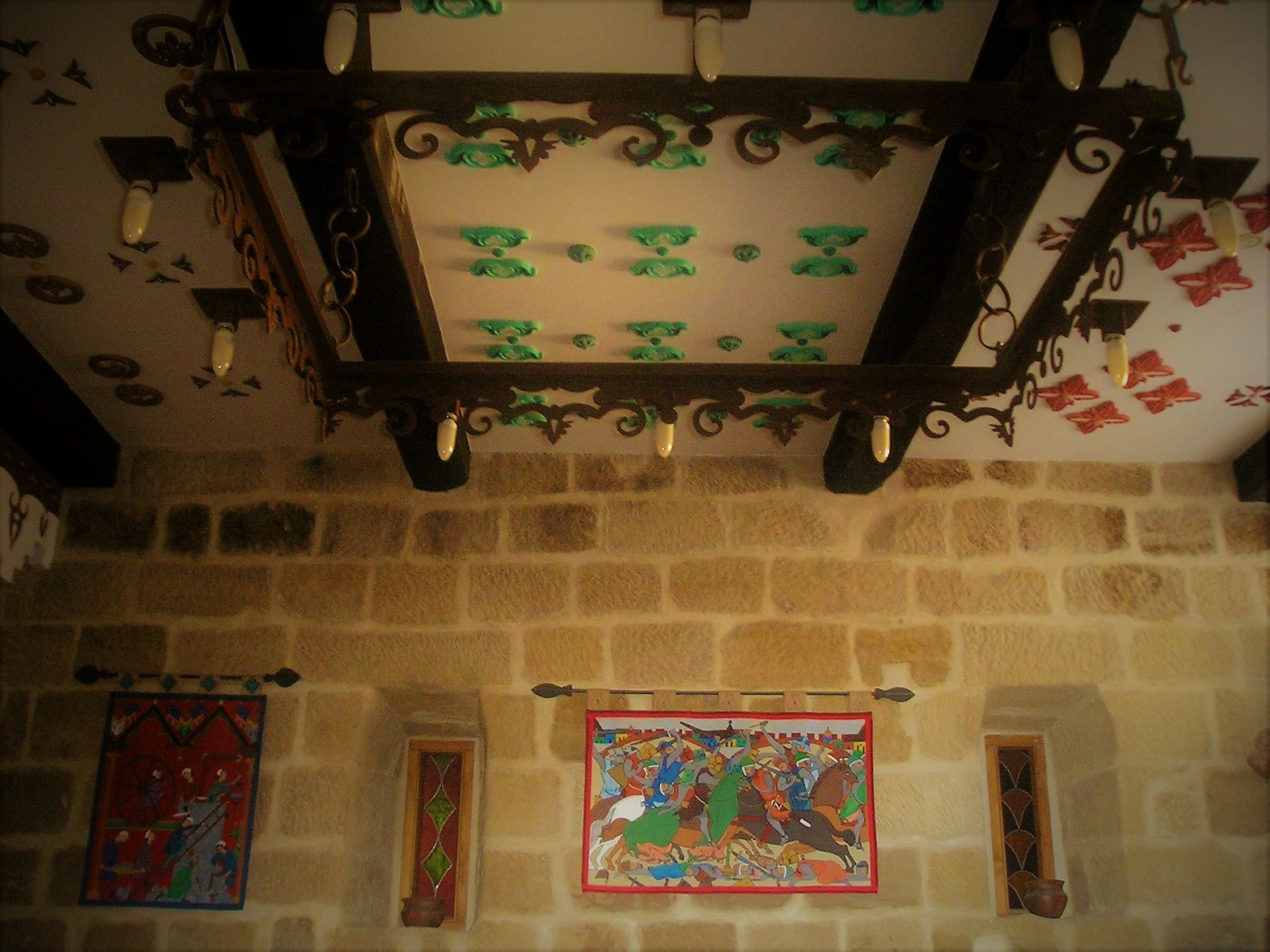 Una habitación con pared de piedra, techo de madera con detalles decorativos y dos ventanas de vitral.