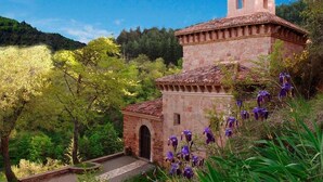 A stone building with a bell tower, surrounded by lush greenery and flowering plants. - TORRE FUERTE S.XIII (Baños de Rioja)