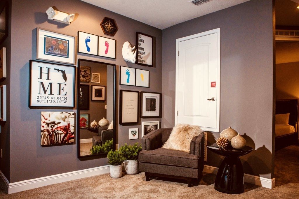 A living room with a grey wall, a large mirror, and various framed pictures. There is a grey armchair with a fur throw and a small table with decorative items.