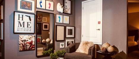 A living room with a grey wall, a large mirror, and various framed pictures. There is a grey armchair with a fur throw and a small table with decorative items.