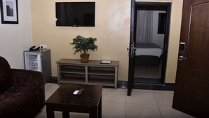 Ein Hotelzimmer mit einem Bett, einem an der Wand montierten Fernseher, einem kleinen Tisch mit Fernbedienung, einer Couch und einer Zimmerpflanze.