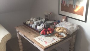 Een hoek van een kamer met een houten tafel waarop appels, koekjes en een theepot staan. Een ingelijst beeld hangt aan de muur boven de tafel.
