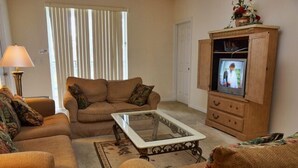 A living room with a sofa set, a glass-top coffee table, a television on a wooden cabinet, and a lamp. - Ahr86 - Sunset Ridge - 5 Bed 4 Baths Villa (Davenport)