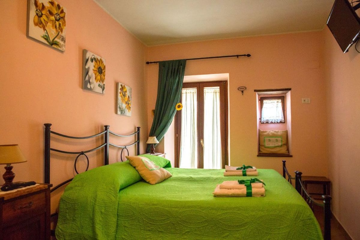 Quarto com colcha verde, cabeceira de madeira, mesa de cabeceira com abajur e janela com cortina.