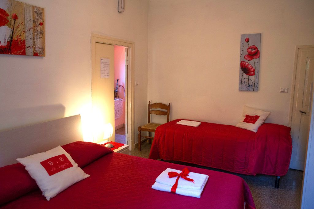 Habitación de hotel con dos camas, cada una con ropa de cama roja y almohadas blancas, una silla de madera y obras de arte en las paredes.