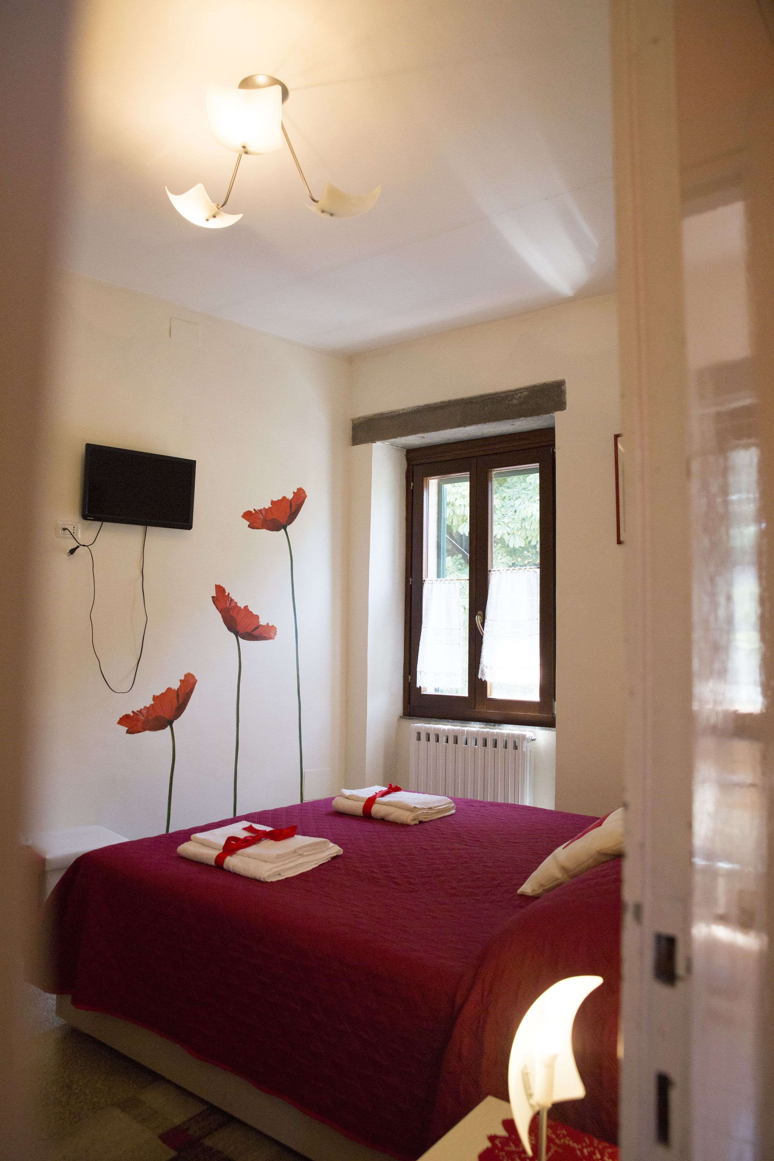 Une chambre à coucher avec une couvre-lit rouge, un téléviseur à écran plat fixé au mur et une fenêtre avec des rideaux.