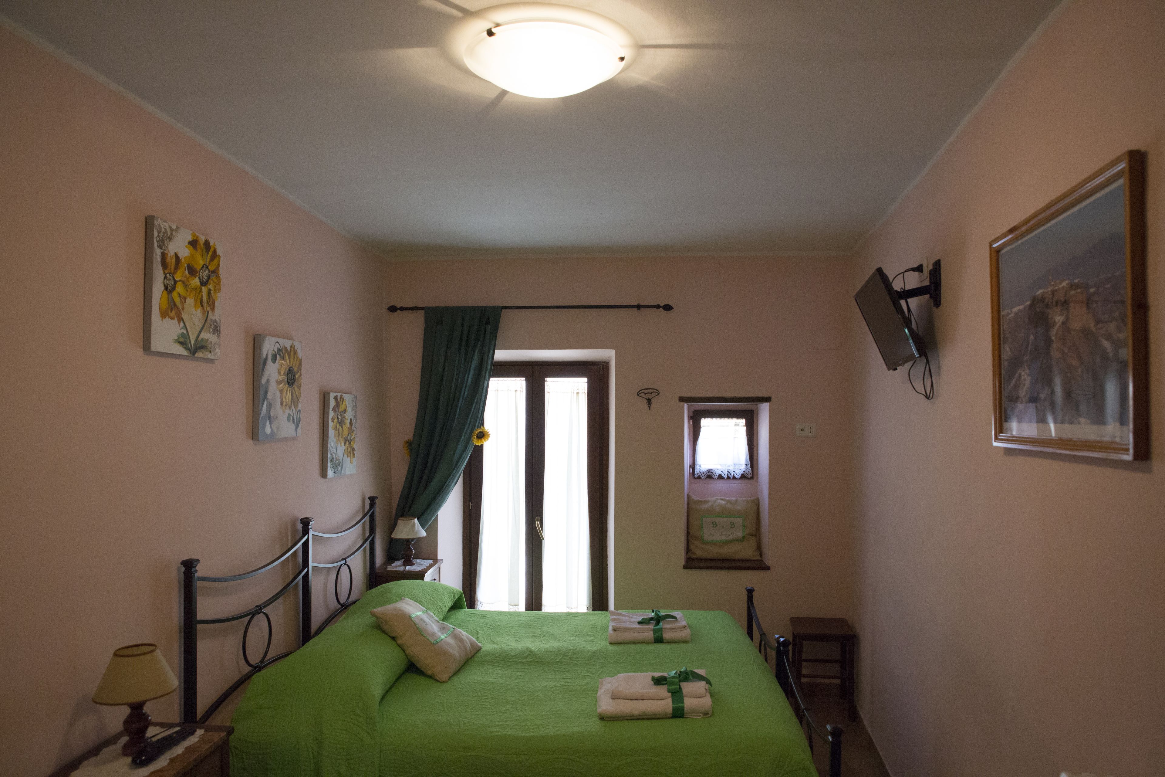 Une chambre avec une couvre-lit verte, une lampe de chevet, un tableau encadré au mur et une fenêtre avec un rideau.