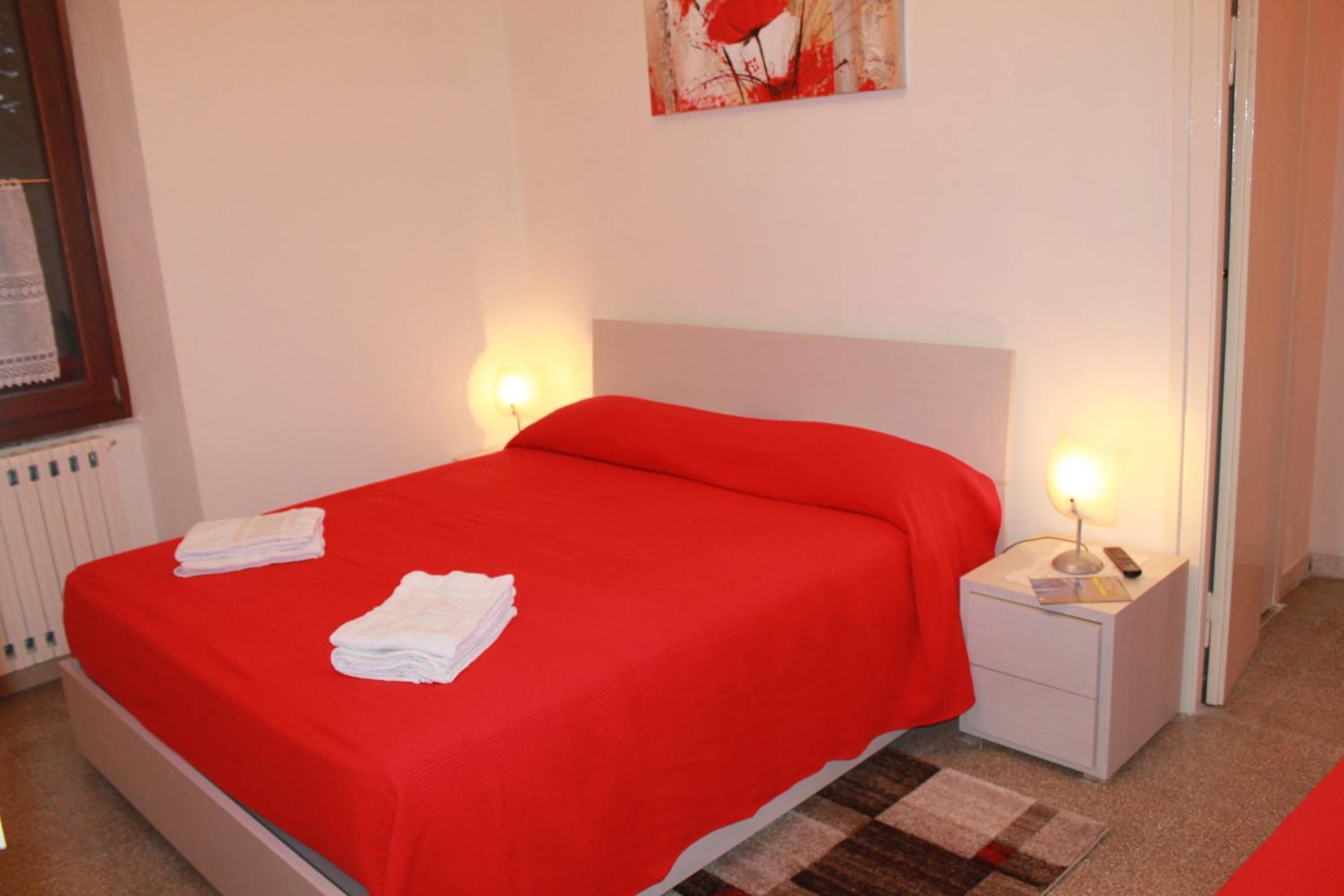 Una habitación de hotel con colcha roja, mesitas de noche blancas y alfombra a cuadros.