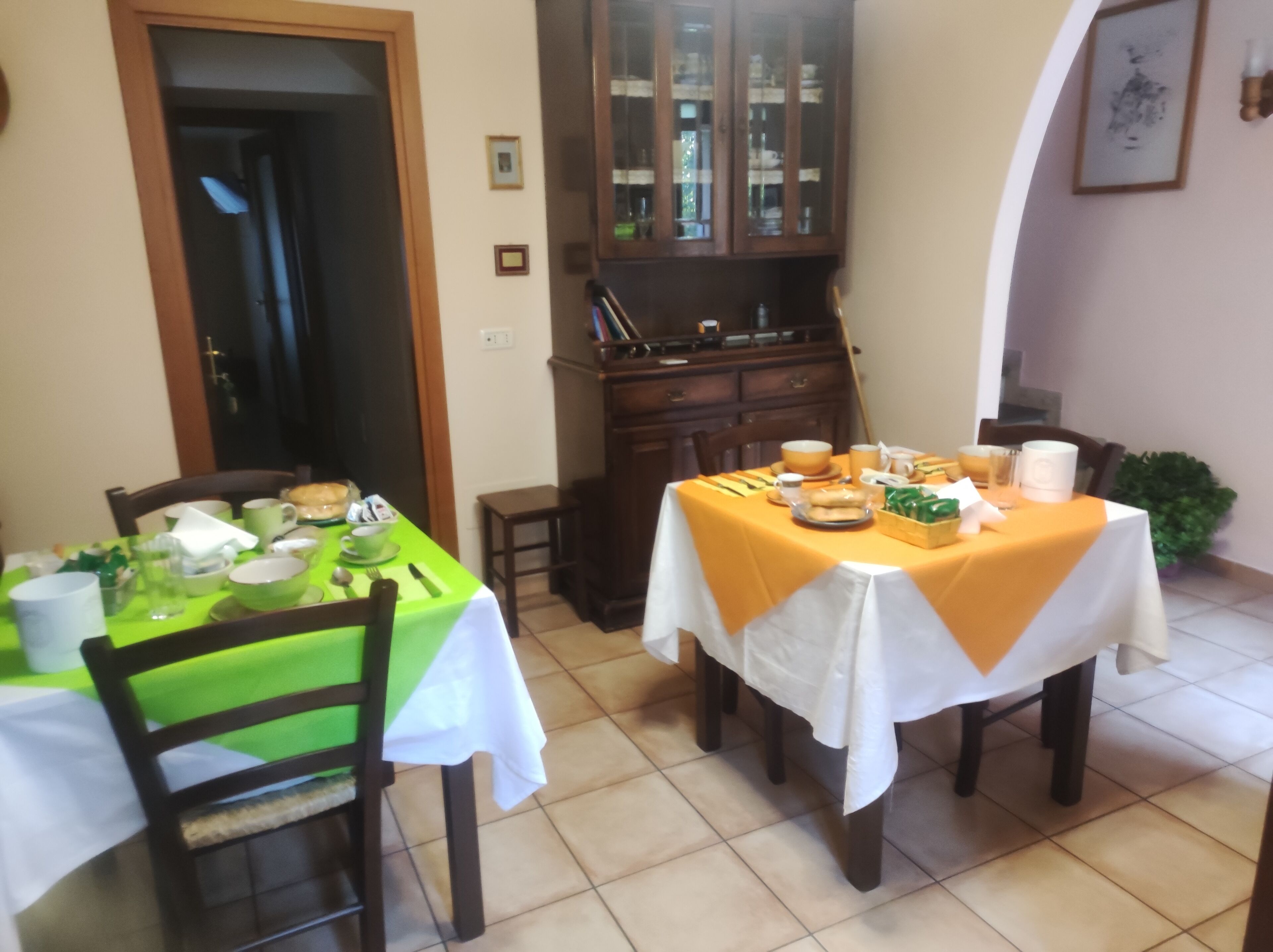 Un coin repas avec deux tables, une verte et une blanche, toutes deux recouvertes de nappes. Des chaises sont disposées autour des tables, et on aperçoit un buffet aux portes vitrées au fond.