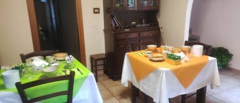 Un coin repas avec deux tables, une verte et une blanche, toutes deux recouvertes de nappes. Des chaises sont disposées autour des tables, et on aperçoit un buffet aux portes vitrées au fond.
