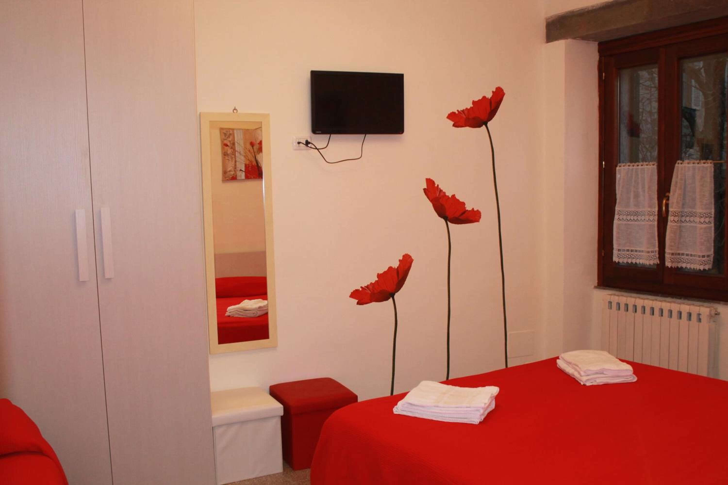 A hotel room with a red bedspread, a mounted TV, a mirror, and red flower decorations.