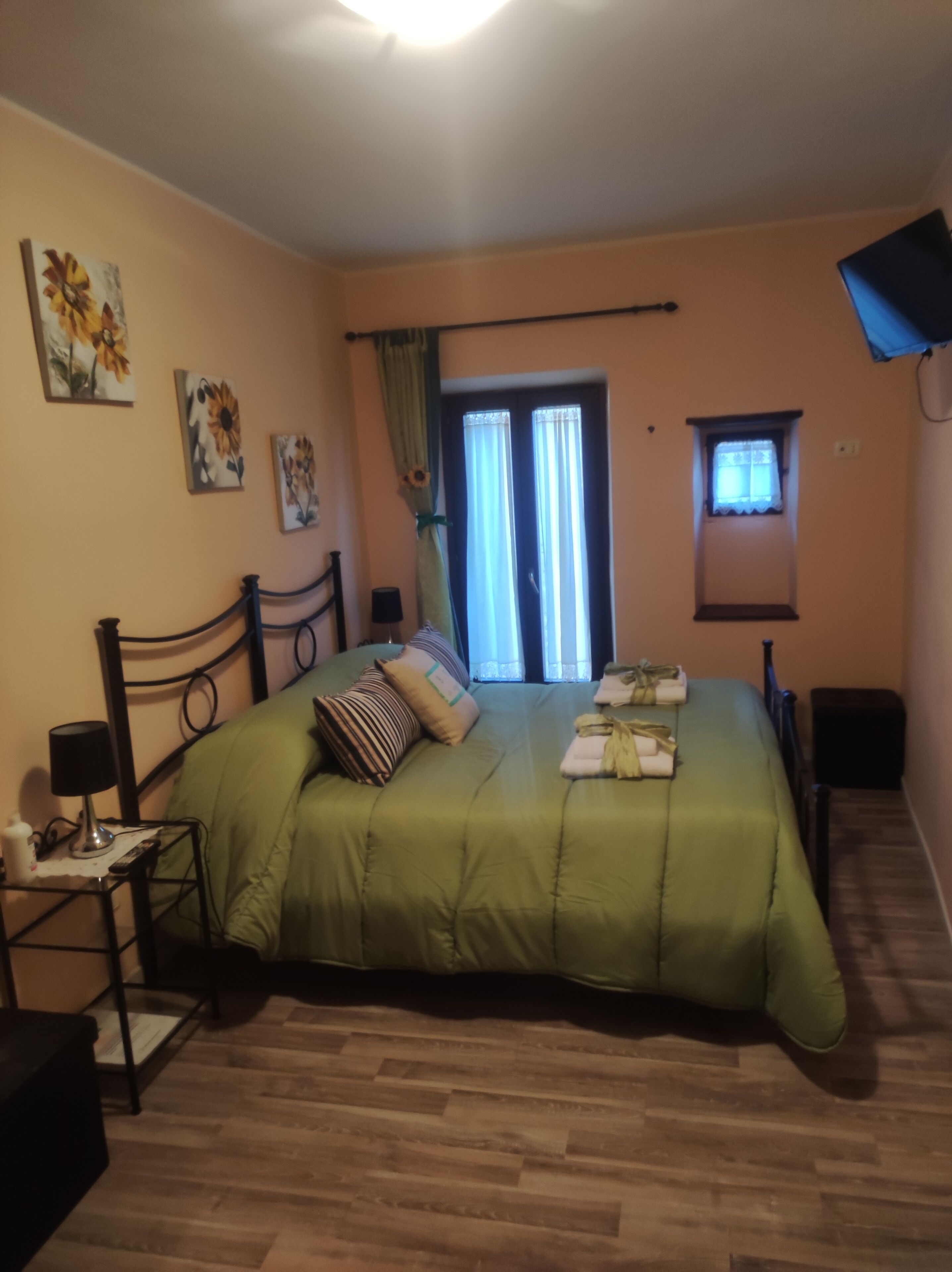 Un dormitorio con una colcha verde, una mesita de noche con lámpara, una ventana pequeña con cortinas y una puerta que da a un balcón.