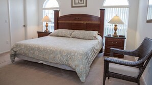 A bedroom with a large bed, two bedside tables with lamps, a wicker chair, and a window with a view of a fence. - Ip60195 - Hampton Lakes - 3 Bed 2 Baths Villa (Davenport)
