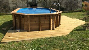 A wooden above-ground swimming pool with a wooden deck, surrounded by a grassy yard and a stone wall. - La Ferme du Grand Launay (Semblancay)