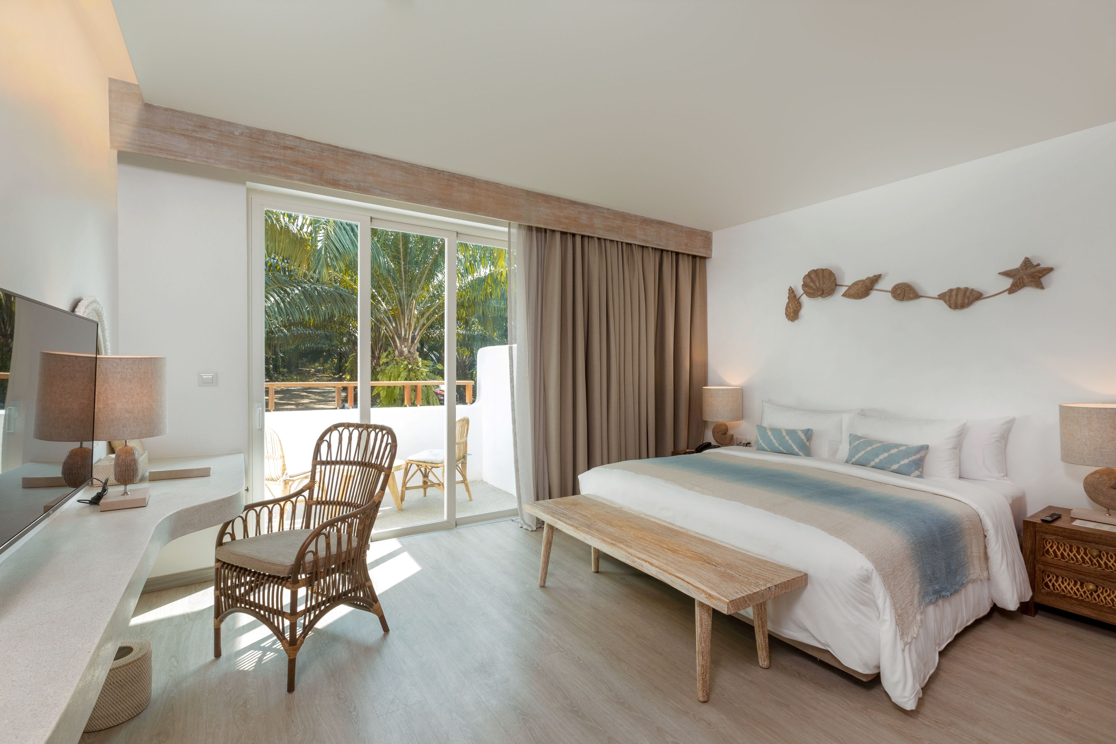 A bedroom with a large bed, a desk, a chair, and a bench. There is a window with curtains, a wall decoration, and a view of greenery outside.