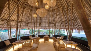 Un espace extérieur aménagé avec des meubles en bambou et un toit de chaume. Des tables et des chaises sont disposées pour les repas.