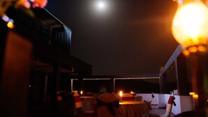 Un área de comedor al aire libre con poca luz, mesas y sillas, una luna en el fondo y un edificio de diseño moderno.