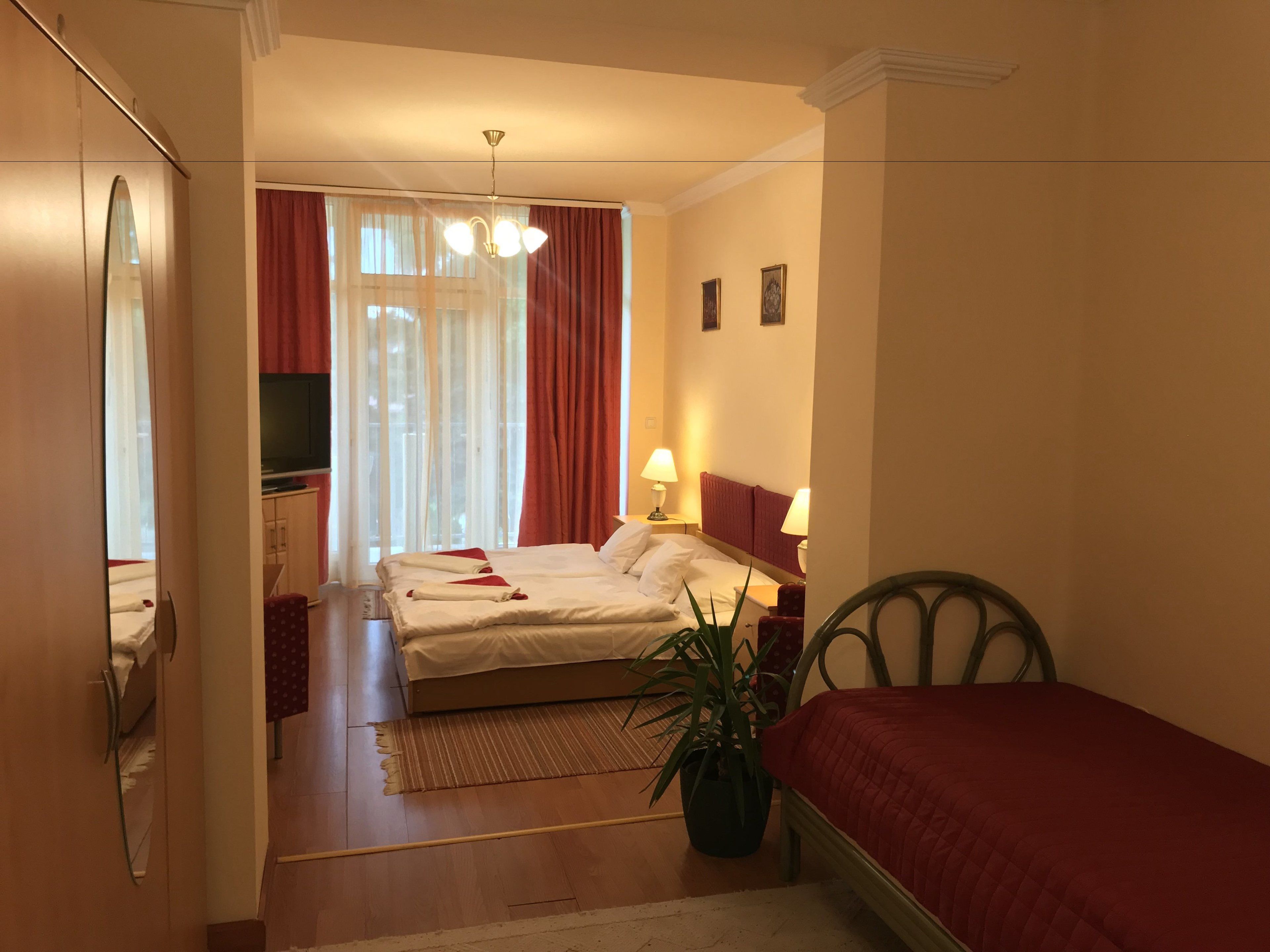 Ein Hotelzimmer mit Bett, Nachttischen, Fernseher, Pflanze und einem Fenster mit Vorhängen.