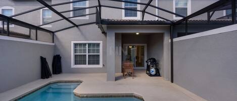 A swimming pool with a covered patio area, a door leading to a covered entryway, and a building in the background.
