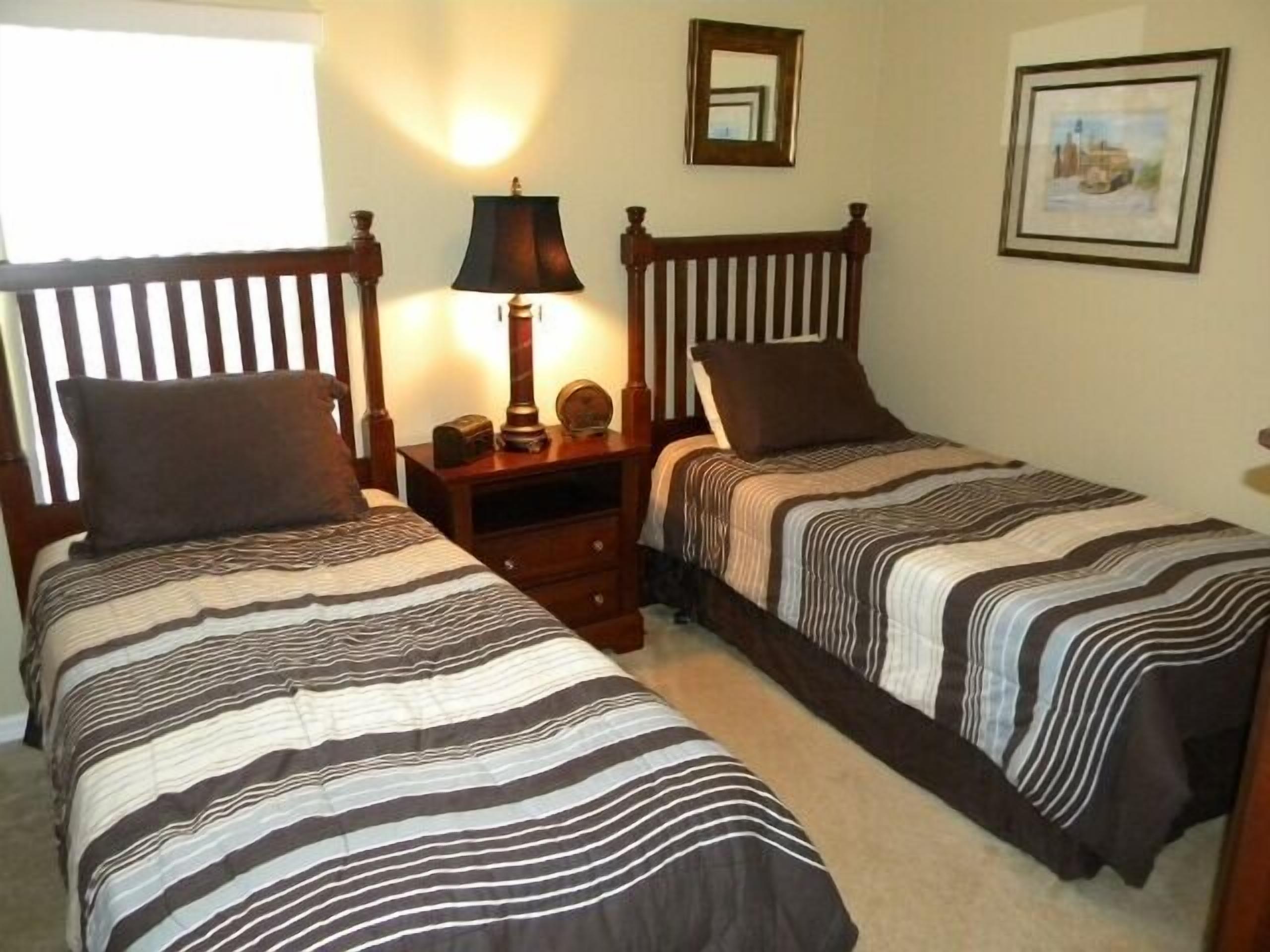 A bedroom with two single beds, each with a striped bedspread, a nightstand with a lamp, a mirror, and a framed picture on the wall.